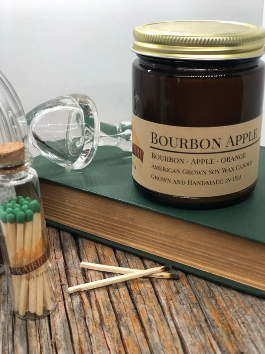 Bourbon apple soy wax candle in amber jar with book, matches, and glass on rustic wood table.