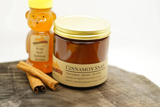 Cinnamon Snap soy wax candle in amber jar with honey bear bottle and cinnamon sticks on wood.