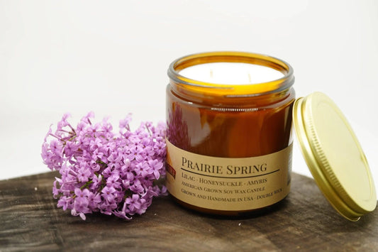 Amber glass soy wax candle labeled Prairie Spring with lilac flowers on wood surface