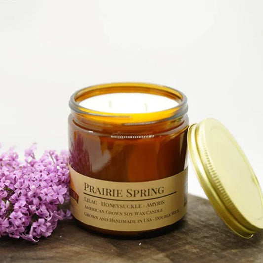 Amber glass jar soy candle labeled Prairie Spring with purple lilacs on a wooden surface