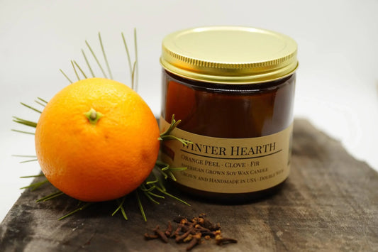 Winter Hearth soy wax candle in amber jar with orange, cloves and fir sprigs on wood surface