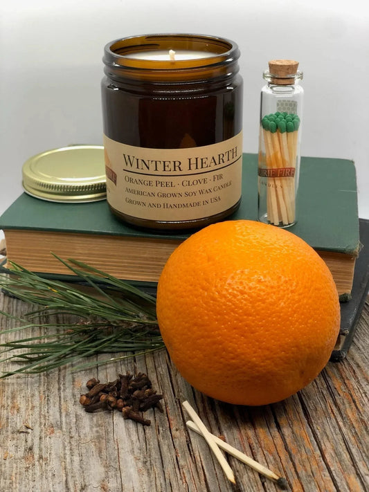 Soy wax candle in amber jar labeled 'Winter Hearth' with orange, clove, fir sprig, and matches