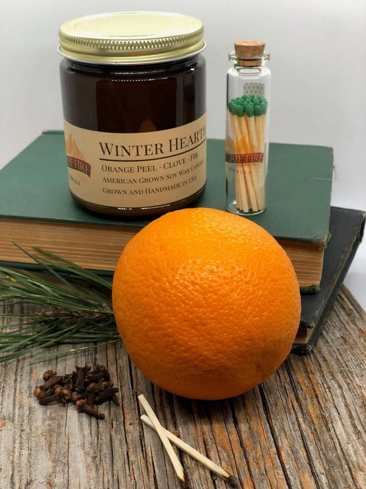 Winter Hearth soy wax candle in amber jar with matches, orange, cloves, and pine on wood surface