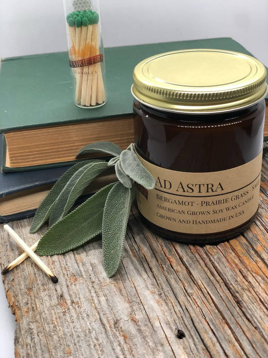 Amber apothecary jar labeled Ad Astra soy wax candle with sage leaves, matches, books, rustic wood surface
