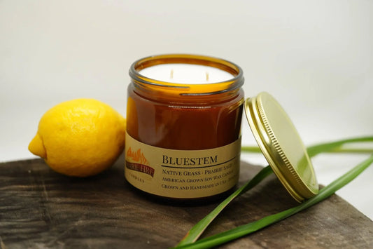 Amber jar soy wax candle labeled Bluestem with lemon and green leaves on wooden surface
