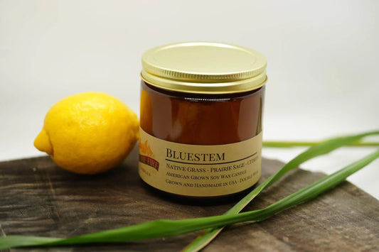Bluestem soy wax candle in amber glass jar with gold lid, lemon, and grass on wood surface