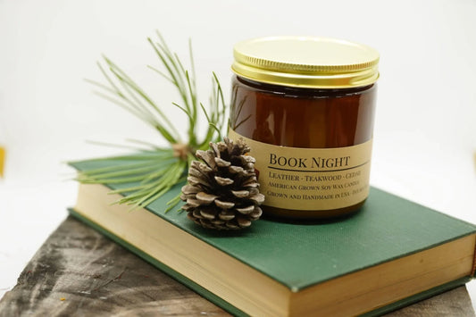 Amber soy wax candle labeled 'Book Night' on green book with pinecone and pine needles