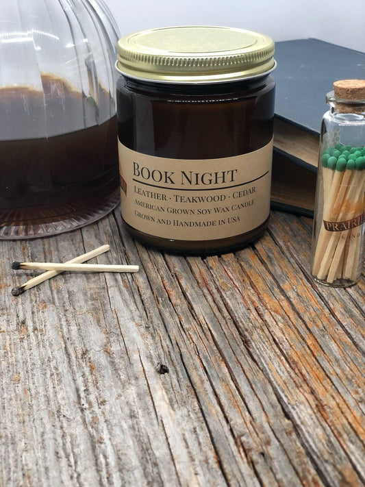 Book Night soy wax candle in amber jar with matches, glass jar, and books on rustic wood table
