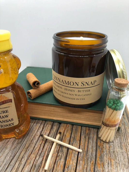 Cinnamon Snap soy wax candle in amber jar with honey, cinnamon sticks, and matches on wood table