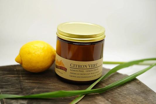 Citron Vert soy wax candle in amber jar with gold lid, fresh lemon and lemongrass on wood.