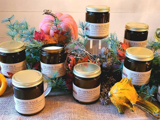 Assorted amber soy wax candle jars with fall scents, surrounded by pine branches and gourds.