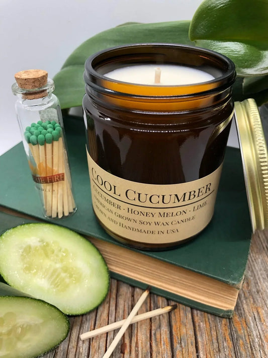 Cool Cucumber soy wax candle in amber jar with sliced cucumber, matches, and green book on wood table