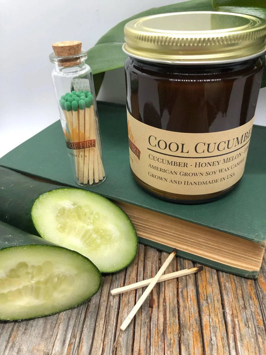 Cool cucumber soy wax candle in amber jar with sliced cucumber, glass jar of matches and green book