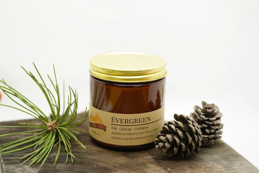 Evergreen soy wax candle in amber jar with gold lid, pine needles and pine cones nearby