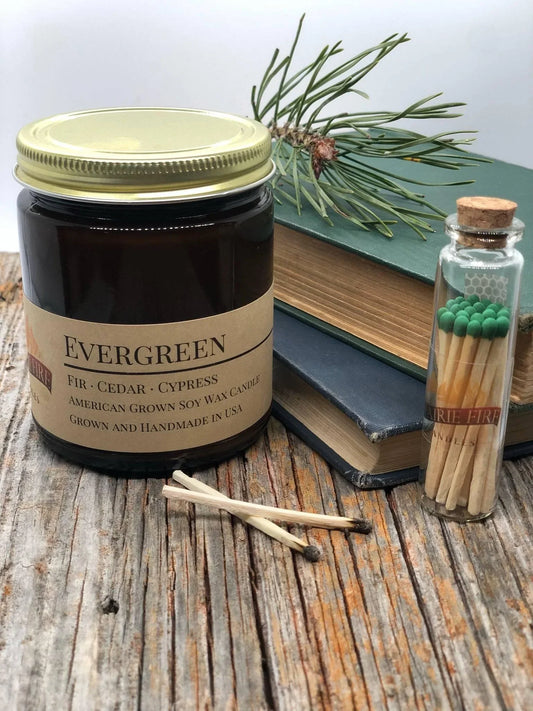 Amber jar evergreen soy wax candle with pine sprig, books, and matches on rustic wood table