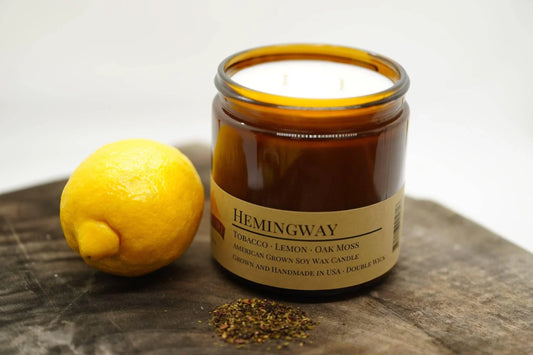 Amber soy wax candle labeled Hemingway with tobacco, lemon, oak moss scents, lemon fruit, and loose herbs on wooden surface