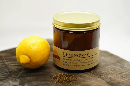 Hemingway soy wax candle in amber jar with gold lid, lemon, and herbs on wooden surface