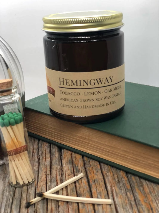 Hemingway scented soy wax candle in amber jar with matches and book on rustic wood surface