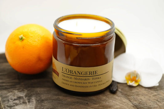 L'Orangerie soy wax candle in amber jar with orange, white orchid, and wood background
