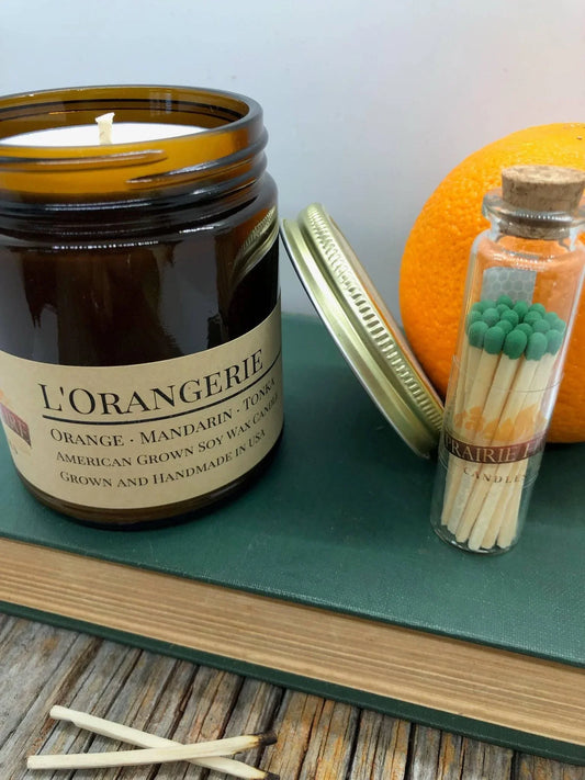 L'Orangerie soy wax candle in amber jar with matches, book, and orange on wood table