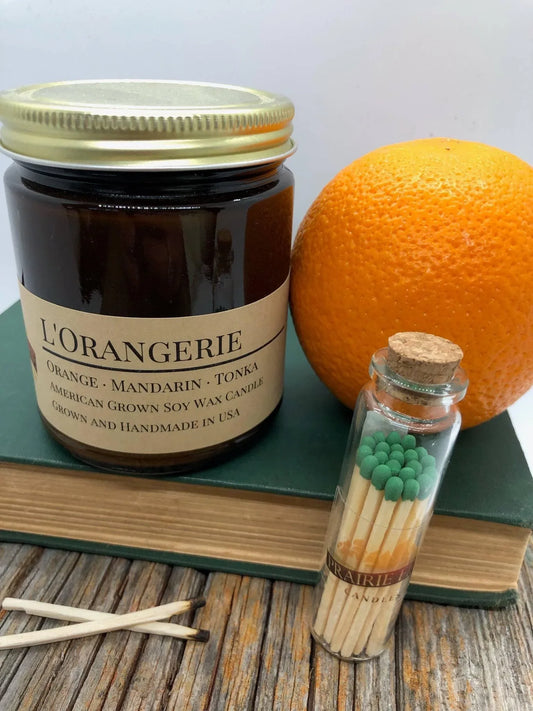 L'Orangerie soy wax candle in amber jar with orange, book, and glass jar matches.