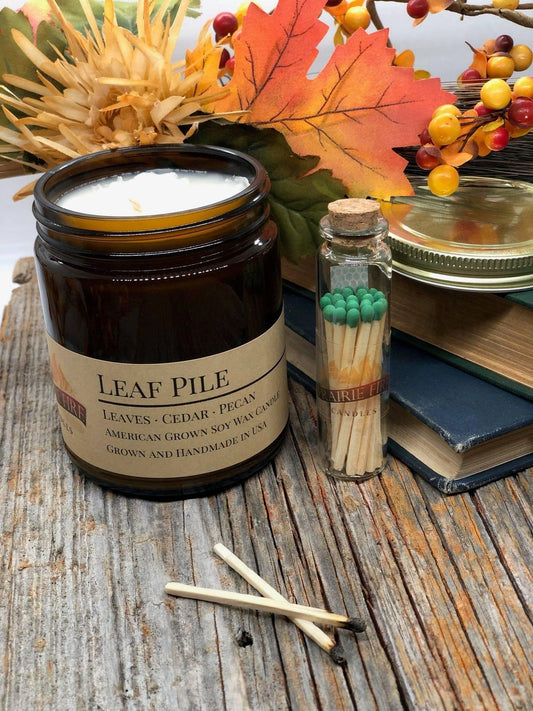 Leaf Pile soy wax candle in amber jar with matches, autumn leaves, and rustic wood background
