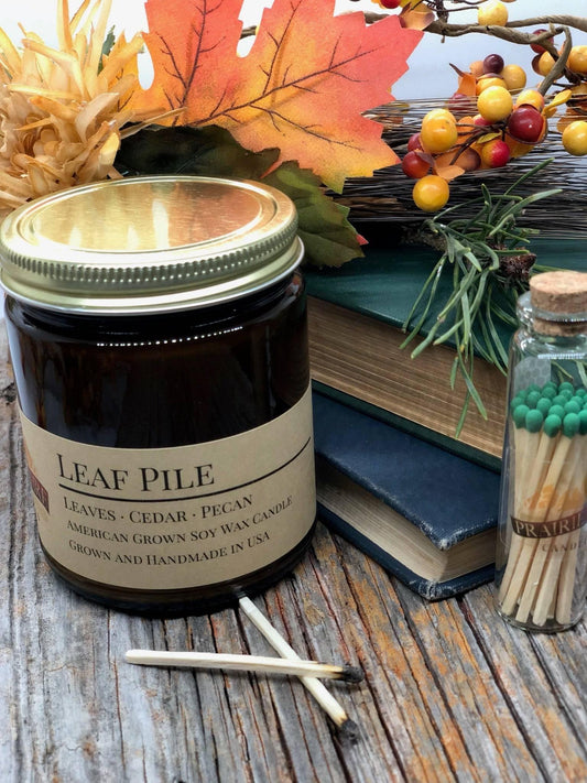Leaf Pile soy wax candle in amber jar with autumn leaves, matches, and rustic wood background