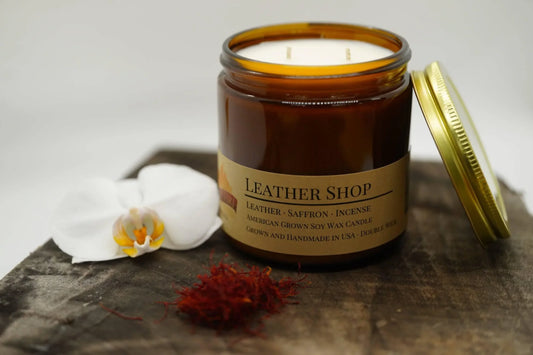 Amber jar soy wax candle labeled Leather Shop, with open lid, white orchid, and saffron on wood