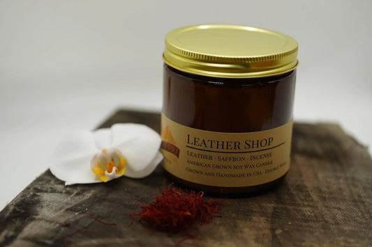 Amber jar soy wax candle labeled Leather Shop with gold lid, white orchid, and saffron accents