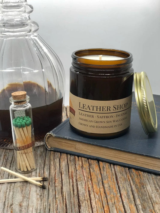 Leather Shop soy wax candle in amber jar on rustic wood with book, matches, and glass bottle.