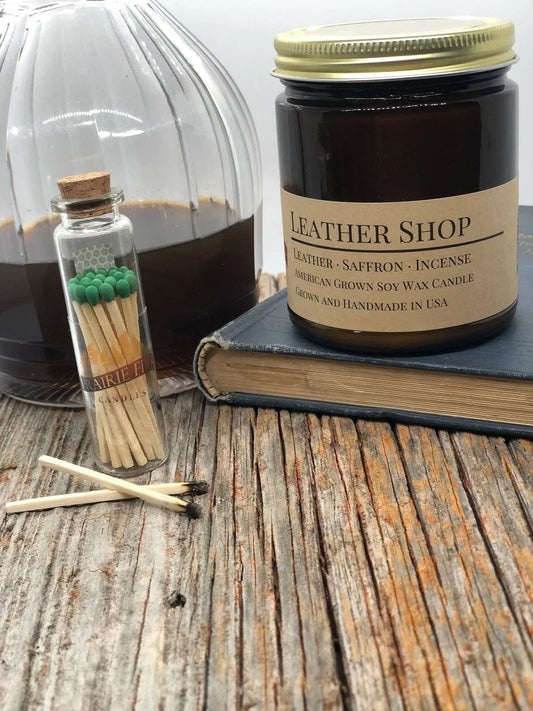 Amber glass soy wax candle labeled 'Leather Shop' with matches on rustic wood table