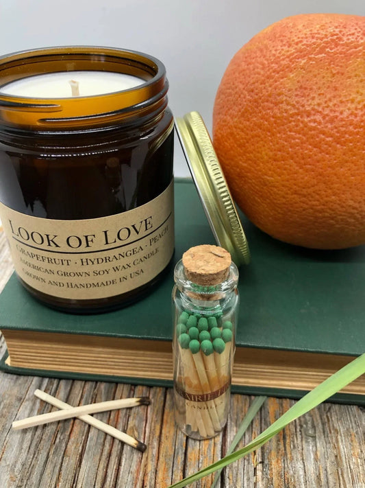 Amber jar soy wax candle labeled 'Look of Love', green-tip matches, grapefruit, and books on wood table