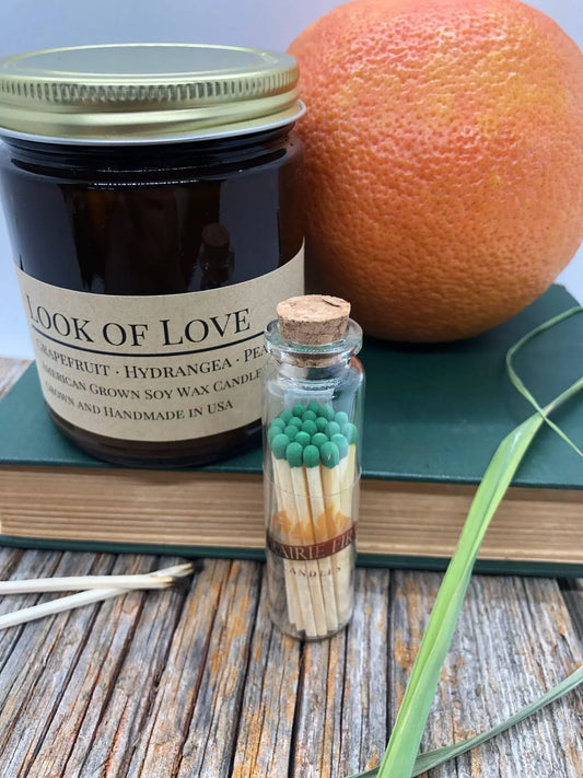 Amber jar soy wax candle labeled 'Look of Love' beside a grapefruit, glass match jar, and books on rustic wood.