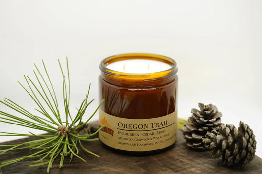 Oregon Trail soy wax candle in amber jar with pine needles and pinecones on wood surface