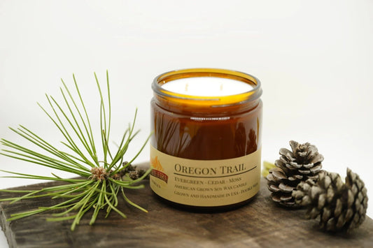 Oregon Trail soy wax candle in amber jar with pine needles and pine cones on rustic wood.