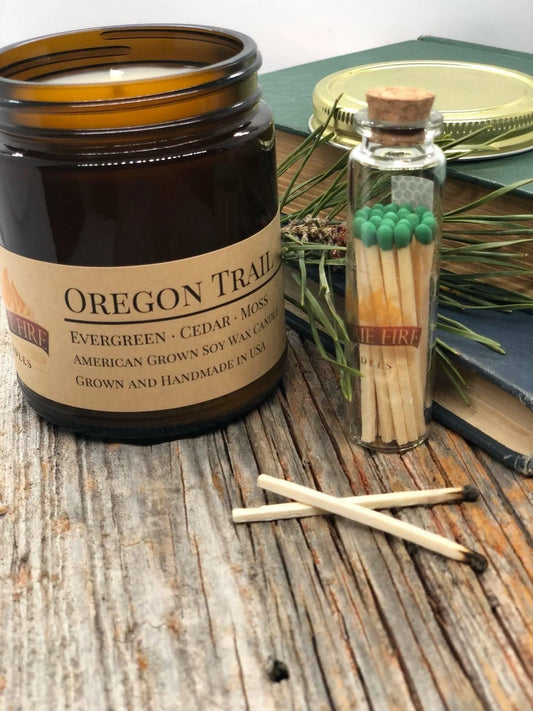 Oregon Trail soy wax candle in amber jar with matches, pine sprig, and books on wood table.