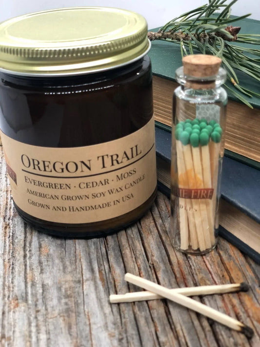 Oregon Trail soy wax candle in amber jar with matches on rustic wood table, pine branch nearby
