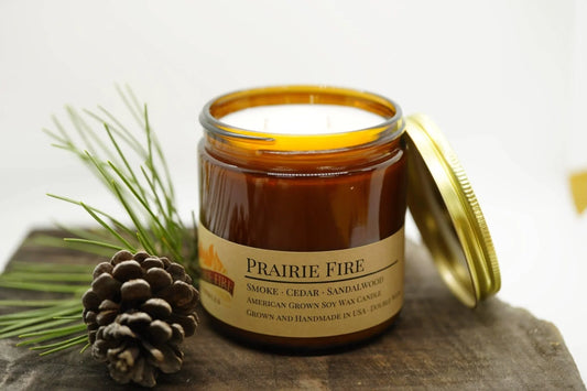 Amber jar soy wax candle labeled Prairie Fire with pine cone and needles on wooden surface