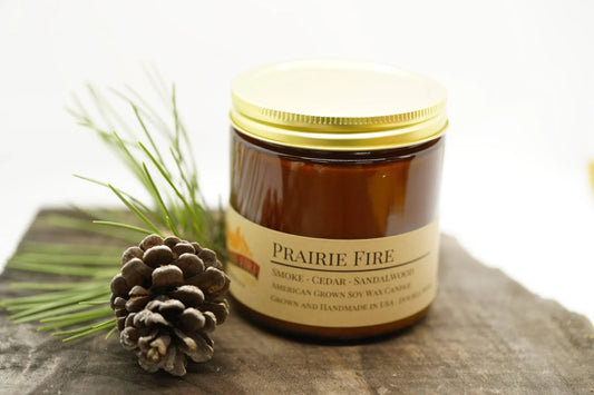 Amber jar soy wax candle labeled Prairie Fire with pinecone and pine needles on rustic wood