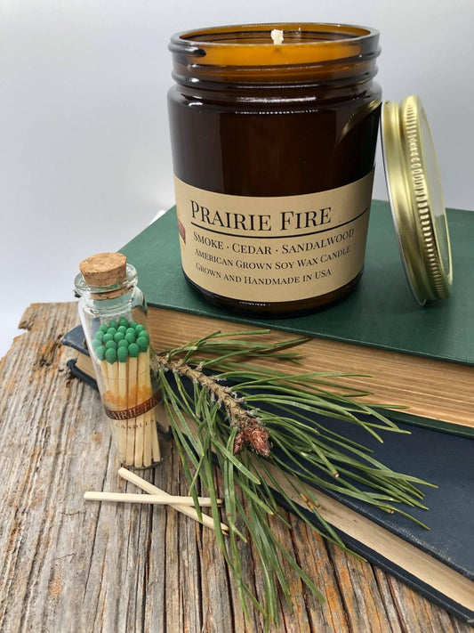 Amber jar soy wax candle labeled Prairie Fire with pine sprig, matches, and books on wood table