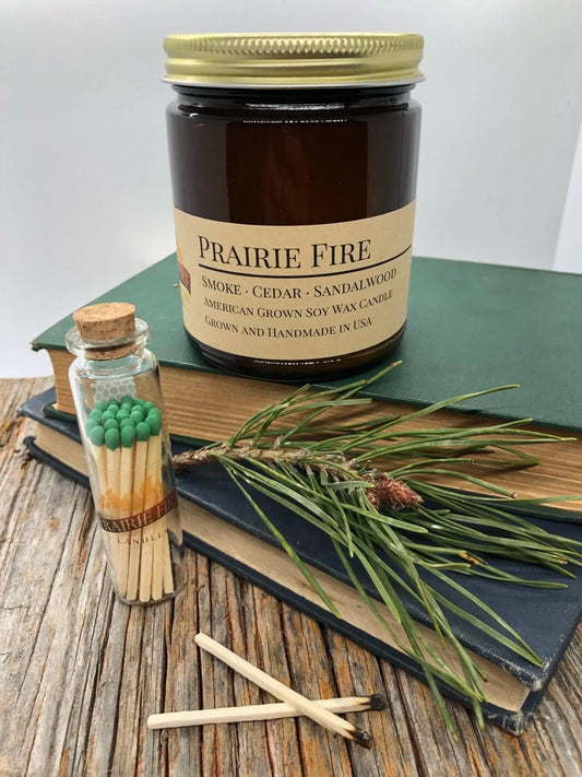 Amber soy wax candle labeled Prairie Fire with matches, pine sprig, and books on wood table