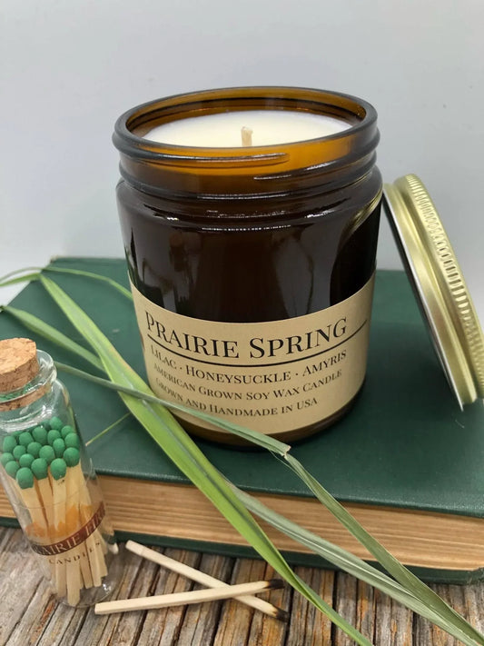Soy wax candle in amber jar labeled Prairie Spring with lilac, honeysuckle, book, and matches