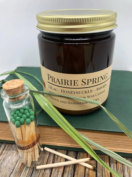 Amber apothecary jar soy wax candle labeled Prairie Spring with lilac, honeysuckle, amyris, next to a glass match jar on book and wood surface.