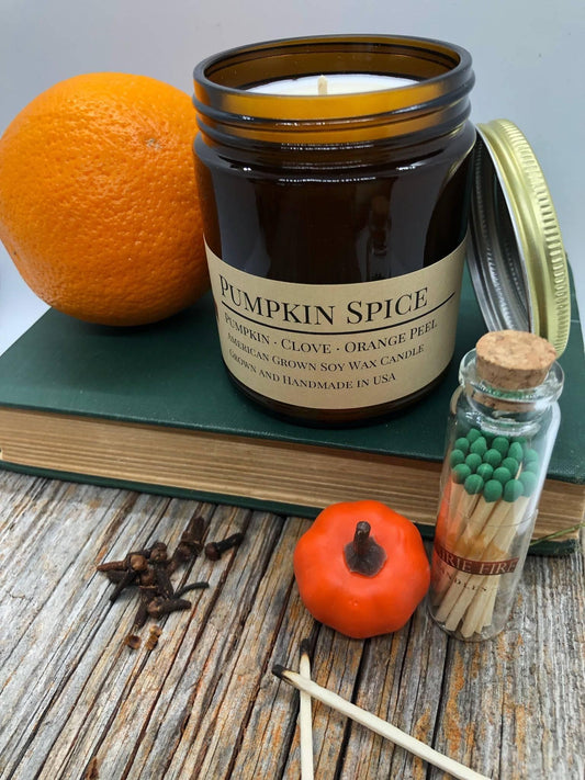 Pumpkin spice soy wax candle in amber jar with orange, cloves, matches, and mini pumpkin on wood table