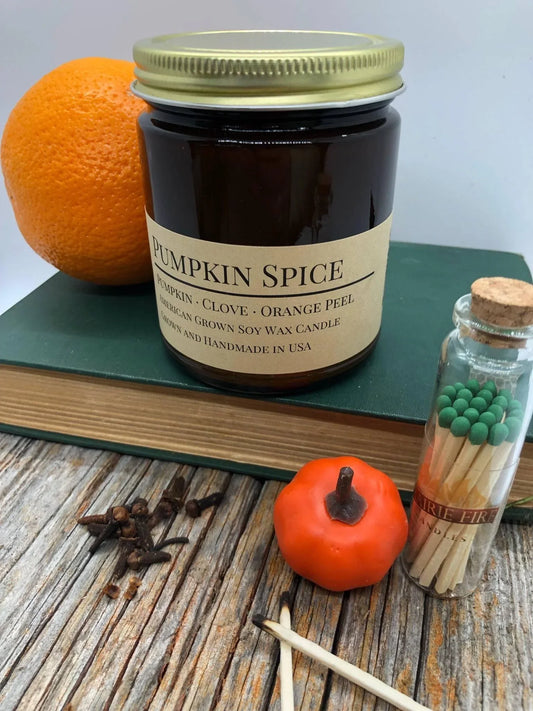 Pumpkin spice soy wax candle in amber jar with orange, small pumpkin, cloves, and matches.