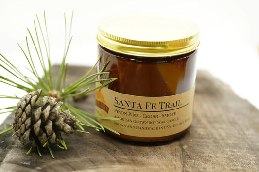 Santa Fe Trail soy wax candle in amber jar with gold lid, pinecone and needles beside it on wood.