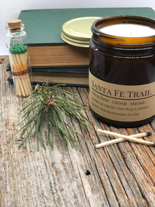 Santa Fe Trail soy wax candle in amber jar with matches, pine sprig, and books on wood surface