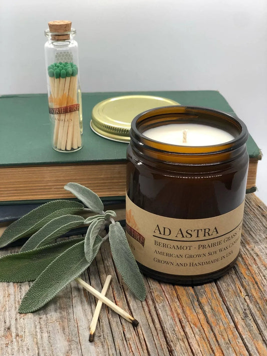 Amber glass soy wax candle labeled 'Ad Astra' with sage leaves, matches, and books on a rustic wood surface.