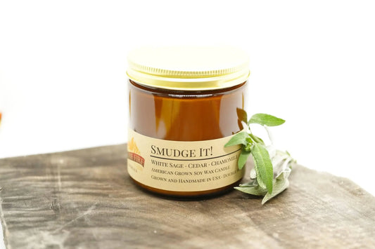 Amber jar soy wax candle labeled Smudge It with sage leaves on wooden surface