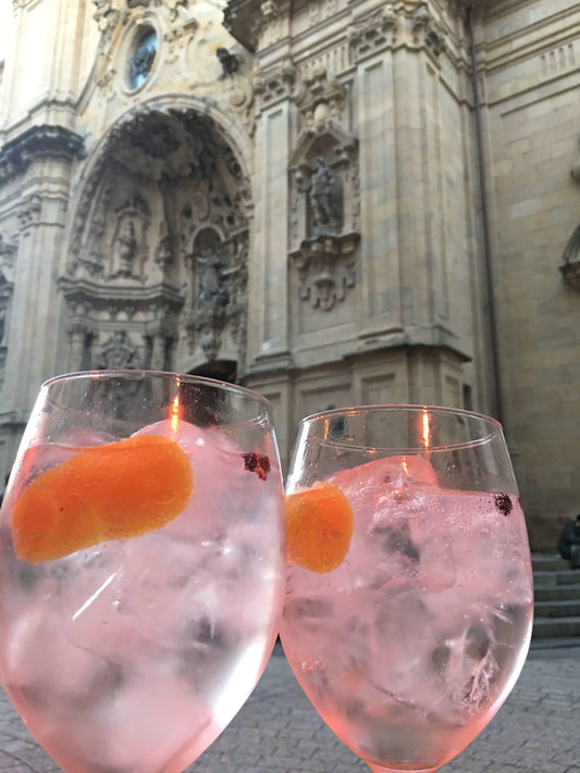 Two Spanish gin and tonic cocktails with orange peel garnish in front of a historic stone building.